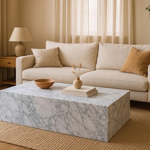 Op de afbeelding: Een witte marmeren salontafel met grijze adering staat op een beige kleed in een woonkamer. De tafel staat voor een witte bank met beige kussens en een bruin kussen.