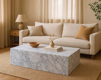 May include: A white marble coffee table with a gray veining pattern sits on a beige rug in a living room. The table is in front of a white sofa with beige cushions and a brown throw pillow.