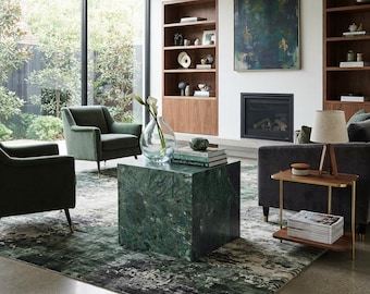 May include: A living room featuring a square green marble coffee table, two dark green velvet armchairs, a dark grey sofa, a two-tier side table with a lamp, a large abstract painting, built-in wooden shelving, and a green and white patterned rug.