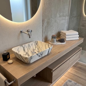 Op de afbeelding: Een moderne badkamermeubel met een rechthoekige wastafel met marmerlook. De wastafel staat op een licht houten kast met lades. Boven de wastafel hangt een ronde spiegel met verlichting. Accessoires: zeepdispenser, kleine vetplant en opgevouwen handdoeken.