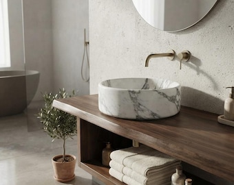 May include: A round white marble vessel sink with grey veining sits atop a dark wood vanity. The vanity features an open shelf holding folded beige towels and small bottles. A wall-mounted brass faucet is positioned above the sink.