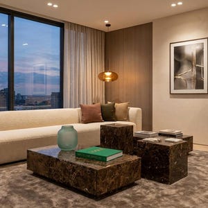 May include: A living room featuring a long cream-colored sofa with three throw pillows, two dark brown marble coffee tables, a green book, a light blue glass vase, a framed abstract print, a potted plant, and a hanging amber pendant light.