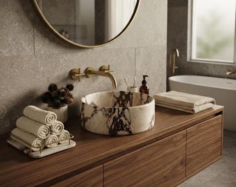 May include: A round marble vessel sink with white and deep purple veining sits on a dark wood vanity. A gold wall-mounted faucet is positioned above the sink. A stack of folded beige towels rests on a marble tray, and a second stack sits nearby.
