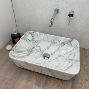Può includere: Un lavabo da bagno in marmo bianco con forma rettangolare e uno scarico al centro. Il lavabo è montato su un pavimento in legno e ha un rubinetto e un dispenser di sapone cromati.