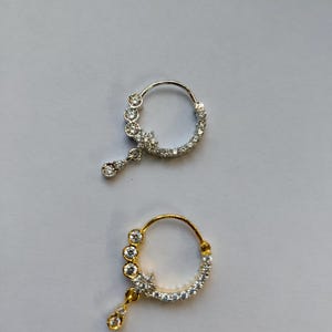 Könnte beinhalten: Zwei Nasenringe, einer silberfarben und einer goldfarben, jeweils mit funkelnden, klaren Edelsteinen verziert. Der silberne Ring hat einen tropfenförmigen Anhänger. Der goldfarbene Ring hat ein ähnliches Design mit passendem Anhänger. Beide sind vor neutralem Hintergrund platziert.