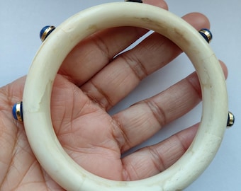 May include: Cream-colored bangle bracelet with gold accents and blue gemstone embellishments. The bracelet is round and appears to be made of a natural material. The gemstones are evenly spaced around the bracelet.