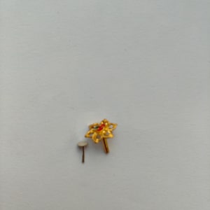 Puede incluir: Un piercing de nariz dorado con un diseño floral y pequeños detalles rojos y verdes. Junto a él, un pequeño poste blanco con la parte superior redonda. La joya está sobre un fondo liso gris claro.