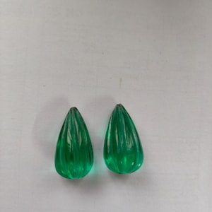 May include: Two teardrop-shaped, emerald green glass beads. Each bead has vertical ridges, giving them a textured appearance. The beads are displayed on a white surface, showcasing their vibrant colour and shape.