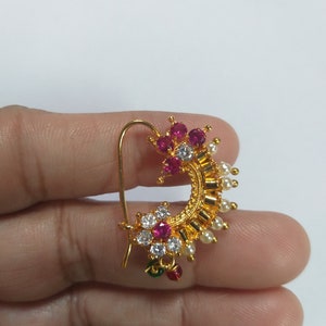 May include: A gold nose ring with a crescent shape and a flower design. The flower has pink and white stones. The ring is decorated with small white pearls.