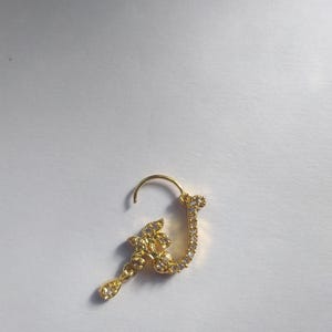 以下が含まれることがあります： 花柄のデザインと、雫型のチャームがついた、金の鼻のリング。リングはクリアなラインストーンで飾られています。