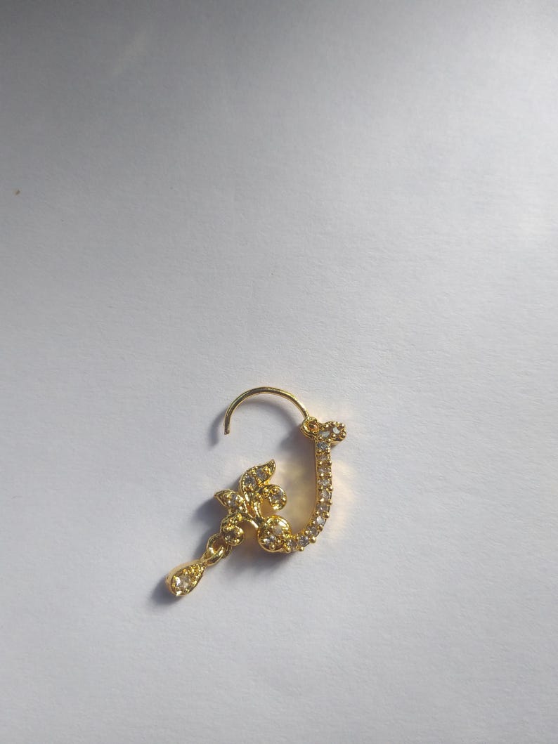 以下が含まれることがあります： 花柄のデザインと、雫型のチャームがついた、金の鼻のリング。リングはクリアなラインストーンで飾られています。