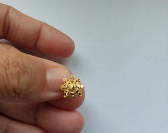May include: A small gold stud earring shaped like a flower with eight petals. The center of the flower features a raised circular detail, and each petal has a textured, faceted surface that reflects light, creating a bright, metallic appearance.