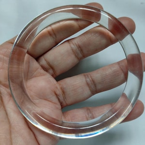 May include: A clear, transparent bangle bracelet. The circular bracelet is held in a hand, showcasing its smooth, polished surface. The light refracts through the clear material, creating subtle highlights and shadows.