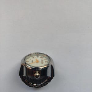 May include: A silver-toned ring watch featuring a round white face with gold-coloured hands and numerals. The watch has a metallic casing and an adjustable, flexible band designed to fit around a finger, combining timekeeping with jewellery.