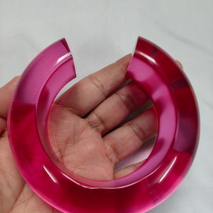 May include: A translucent, magenta-colored bangle bracelet with a split design. The bracelet is a thick, curved band, showcasing a glossy finish. The open design allows for easy wear. The bracelet is held against a white background.