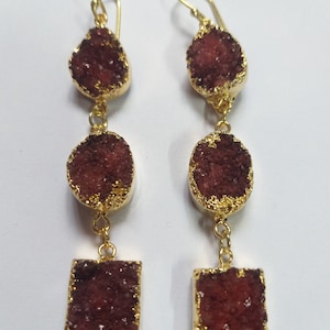 Puede incluir: Un par de pendientes largos de oro con tres piedras rojas de cristal en cada pendiente. Las piedras tienen forma de lágrima, ovalada y rectangular.