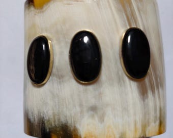 Puede incluir: Un brazalete ancho y curvo hecho de cuerno, con un patrón natural crema y marrón. Tres piedras negras ovaladas están engastadas en el brazalete con biseles de metal dorado. El brazalete tiene un acabado pulido.