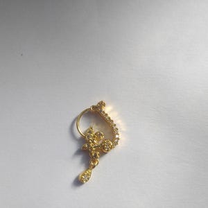 以下が含まれることがあります： 花柄のデザインとクリアなラインストーンが付いた金の鼻のリング。リングはフープの形で、ぶら下がっているドロップがあります。