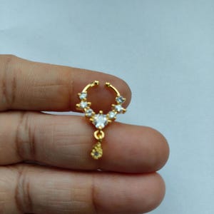 以下が含まれることがあります： 金色の三日月型のノーズリング。透明でキラキラ光る宝石で飾られています。中央からは涙型のチャームがぶら下がっています。ジュエリーは明るい背景に保持されています。