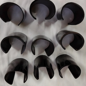 Könnte beinhalten: Neun schwarze, gebogene Armreifen mit offenem Ende sind auf einer weißen Oberfläche angeordnet. Die Armreifen haben eine glänzende Oberfläche und ein schlichtes, modernes Design. Die gebogene Form deutet darauf hin, dass sie für das Handgelenk bestimmt sind.