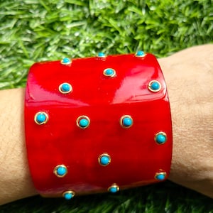 May include: A wide, glossy red cuff bracelet featuring scattered, small, round turquoise-colored cabochons, each individually set within a thin, circular gold-toned metal frame, creating a distinct contrast against the vibrant red surface.