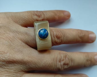 以下が含まれることがあります： 中央に青い宝石がセットされたベージュの四角いリング