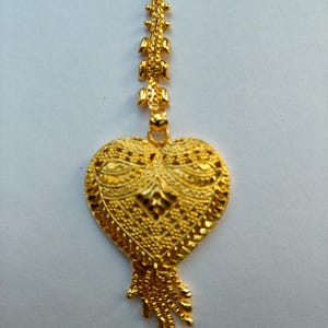 Op de afbeelding: Een goudkleurige hanger met een hartvormig ontwerp. De hanger heeft ingewikkelde details en een fijne ketting. Het hart heeft een getextureerd oppervlak en een kleine kwast aan de onderkant. De sieraden zijn waarschijnlijk van goud.