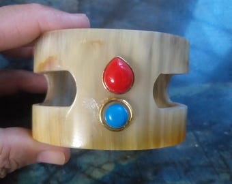 May include: A wide, beige-colored cuff bracelet with two cut-out sections. The bracelet is adorned with a red gemstone and a blue gemstone, both set in gold-colored metal. The bracelet is held by a hand.