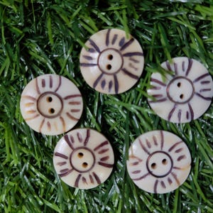 Peut inclure: Cinq boutons ronds, blanc cassé, avec un motif de soleil en marron foncé. Chaque bouton a deux trous pour la couture. Les boutons sont disposés sur un lit d'herbe artificielle verte.