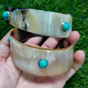 Op de afbeelding: Twee brede armbanden gemaakt van gepolijst, doorschijnend hoornmateriaal met natuurlijke crème, beige en donkerbruine strepen. Elke armband is voorzien van ronde, helder turquoise cabochonstenen in goudkleurige metalen zettingen.