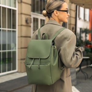 Könnte beinhalten: Ein grüner Rucksack mit Klappenverschluss und Kordelzugdetail. Der Rucksack hat verstellbare Träger und eine Vordertasche. Die Tasche besteht aus einem glatten Material und wird im Freien getragen.