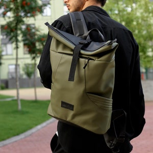 Puede incluir: Mochila verde oliva con solapa superior y correas negras. La bolsa tiene un bolsillo frontal y una pequeña etiqueta negra. La persona de la imagen lleva una camisa negra y gafas de sol.