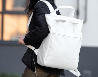 Può includere: Uno zaino bianco con maniglia superiore e spallacci regolabili. Lo zaino ha una tasca frontale con cerniera e un design minimalista. La borsa è realizzata in materiale liscio. Lo zaino è indossato.
