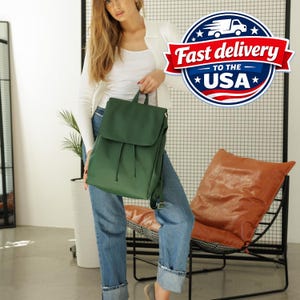 May include: A forest green backpack with a fold-over flap and drawstring closure. The bag features a top handle and adjustable shoulder straps. The design is simple and functional, showcasing a smooth matte finish and a structured rectangular shape.