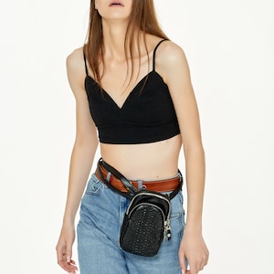 May include: A black crop top with thin straps and a V-neck is worn with light blue jeans and a brown belt. A small, black, textured fanny pack with a zip is worn around the waist. The outfit is simple and stylish.