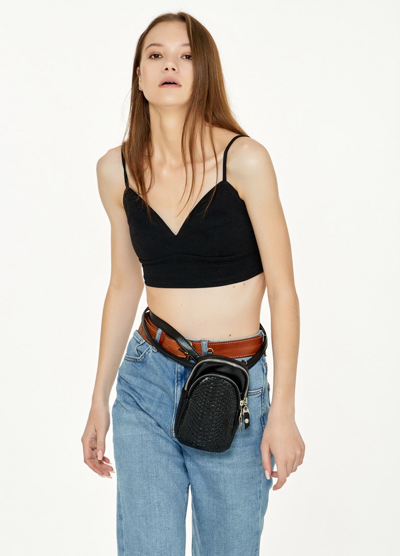 May include: A black crop top with thin straps and a V-neck is worn with light blue jeans and a brown belt. A small, black, textured fanny pack with a zip is worn around the waist. The outfit is simple and stylish.