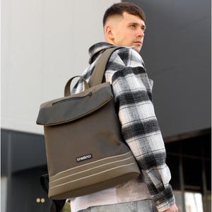 Puede incluir: Mochila verde oliva con cierre de solapa y asa superior. La mochila tiene un cuerpo texturizado con rayas blancas y el nombre de la marca "SAMBAG" en la parte delantera. La persona lleva una camisa a cuadros en blanco y negro.