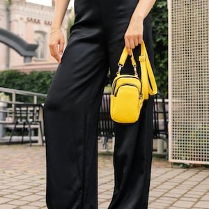 May include: A person wearing black wide-leg trousers and sandals holds a small, bright yellow crossbody bag. The bag has a rectangular shape with a rounded top and a black zip. The background includes a brick walkway and a building.