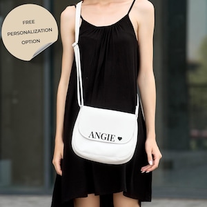 Op de afbeelding: Witte crossbodytas met de naam "ANGIE" en een hartsymbool op de voorkant. De tas heeft een klepsluiting en een witte riem. Een sticker linksboven leest "FREE PERSONALIZATION OPTION".