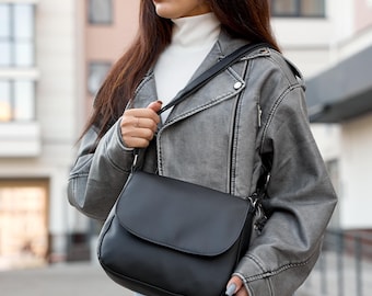Pode incluir: Uma bolsa preta transversal com fecho de aba. A bolsa é transportada por uma alça preta e é combinada com uma jaqueta cinza e jeans azuis. A bolsa é um estilo clássico, adequado para o uso diário.