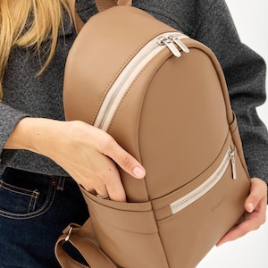 Op de afbeelding: Een effen beige rugzak met een gladde, matte afwerking, een hoofdvak met ritssluiting en twee zilverkleurige trekkers, een kleiner voorvak met ritssluiting en verstelbare schouderbanden. De tas is voorzien van zijvakken en een subtiel logo.