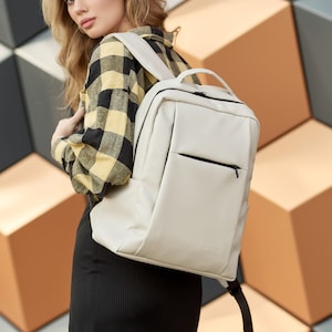 Op de afbeelding: Een lichtbeige rugzak met een voorvak en een handvat aan de bovenkant. De rugzak heeft een minimalistisch ontwerp en een zwarte ritssluiting. De merknaam "SunBag" is rechtsonder zichtbaar. De rugzak wordt gedragen.