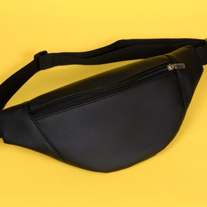 May include: Black fanny pack with a zippered main compartment and adjustable black strap. The bag is made of a smooth, leather-like material and is set against a yellow background.