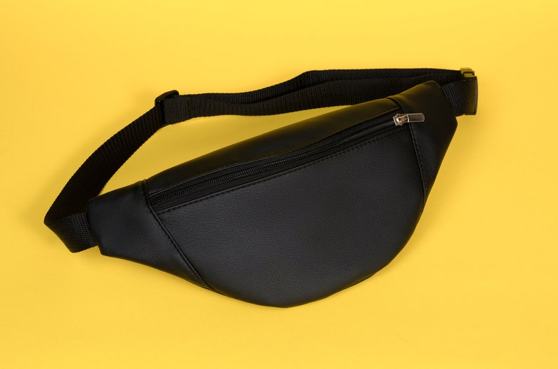May include: Black fanny pack with a zippered main compartment and adjustable black strap. The bag is made of a smooth, leather-like material and is set against a yellow background.