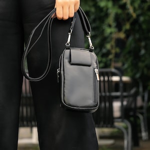May include: A black crossbody bag with a rectangular design and a small front pocket. It features a zip closure and a long, adjustable strap. The bag is made of a smooth, leather-like material.