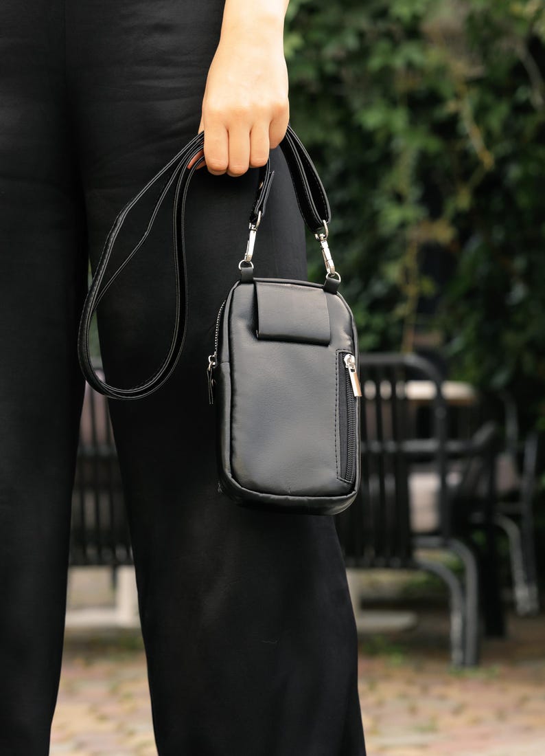 May include: A black crossbody bag with a rectangular design and a small front pocket. It features a zip closure and a long, adjustable strap. The bag is made of a smooth, leather-like material.
