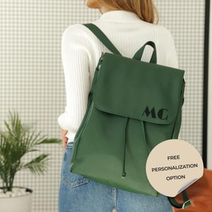 Könnte beinhalten: Ein dunkelgrüner Rucksack mit Klappenverschluss und Kordelzug. Auf der Vorderseite sind die Initialen "MC" aufgedruckt. Ein Aufkleber unten rechts lautet "FREE PERSONALIZATION OPTION".