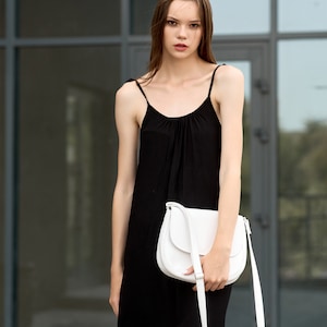 Pu&ograve; includere: Abito sottoveste nero con sottili spalline e borsa a tracolla bianca curva. La borsa ha una lunga tracolla e una fibbia argentata. L'abito &egrave; al ginocchio e la modella &egrave; in piedi davanti a un edificio di vetro.