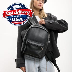 May include: A black backpack with a textured finish, featuring a main zippered compartment and a smaller front zippered pocket. The bag includes adjustable black shoulder straps and silver-toned metal zipper pulls, presented in a clean design.