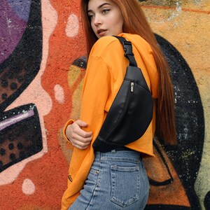 May include: A black fanny pack with a zippered pocket and adjustable strap. The bag is worn across the body. The person is wearing an orange hoodie and blue jeans. The background is a colorful graffiti wall.